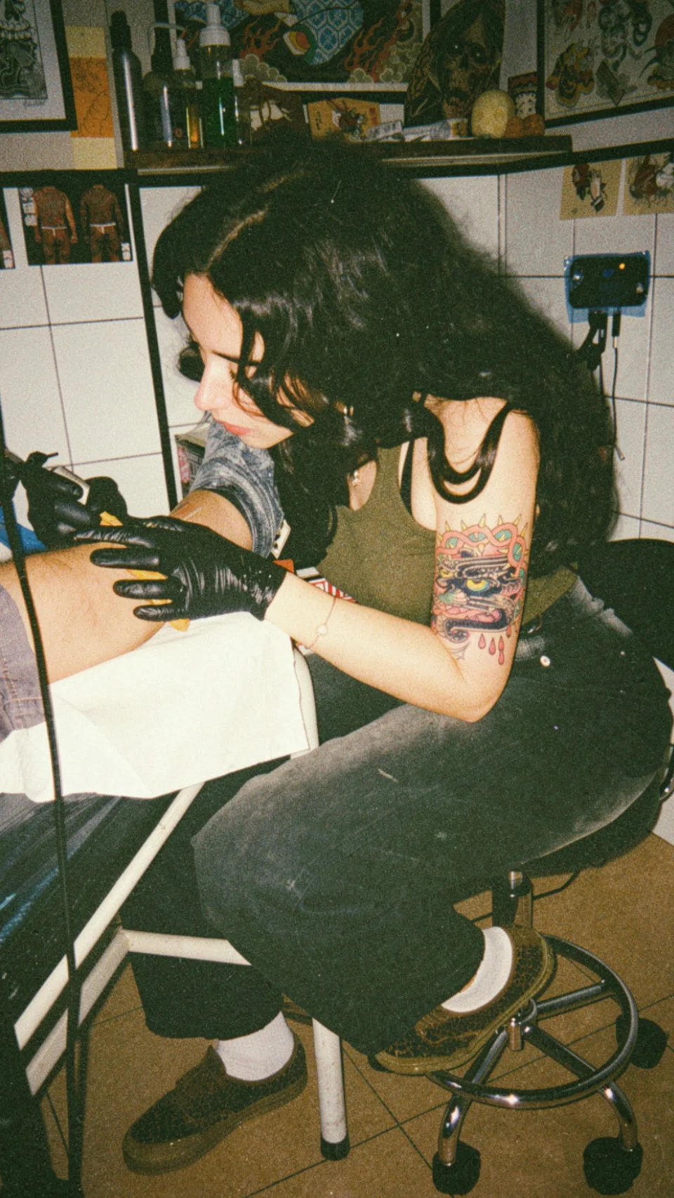 PekoBoop tatuando un brazo en su estudio, rodeada de láminas de flash tradicional.